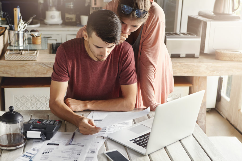 Young family managing budget, reviewing their bank accounts using generic laptop pc and calculator in kitchen. Husband and wife doing paperwork together, paying taxes online on notebook computer